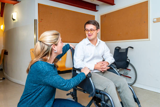 A support worker kneeling to talk with a young man seated in his wheelchair